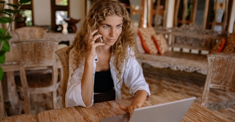 A blonde woman looking shocked while using a laptop while she holding a smartphone to her ear